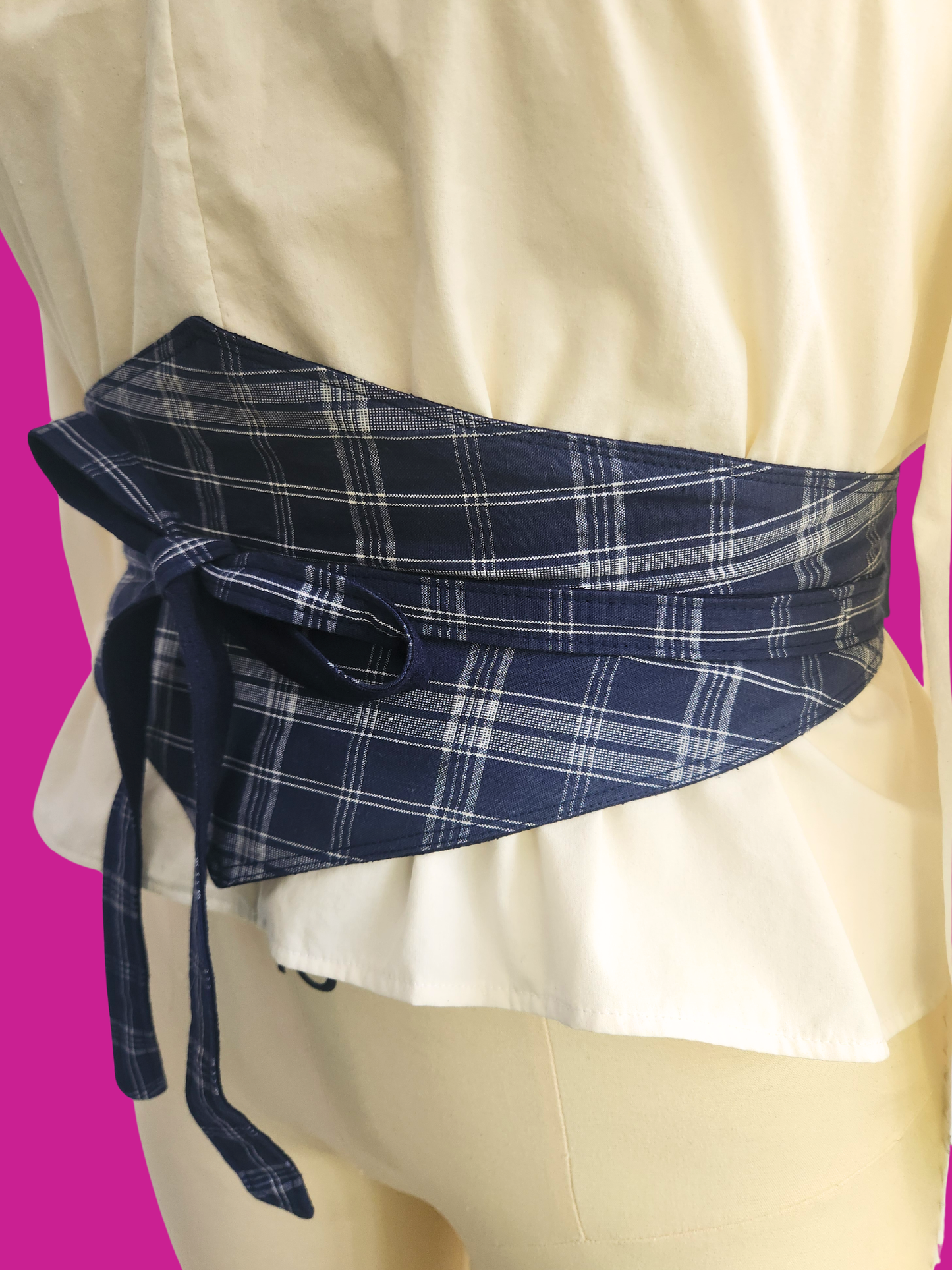Blue plaid belt tied around a person wearing a white dress on a pink background