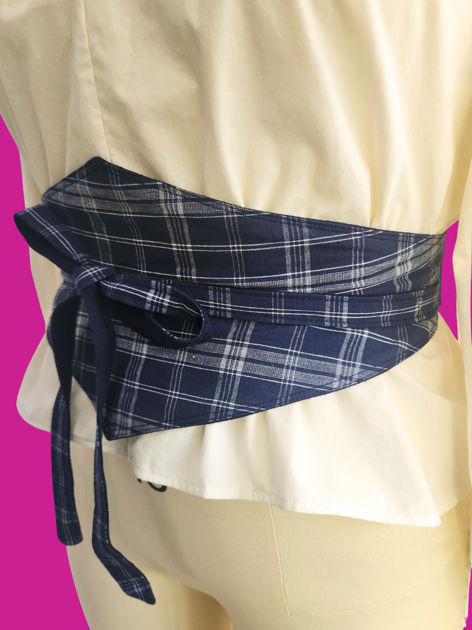 Blue plaid belt tied around a person wearing a white dress on a pink background